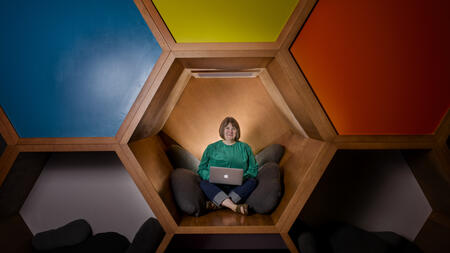 Kristin Fontichiaro sitting cross-legged in colorful cubes at a library, wearing a green sweater and smiling. 