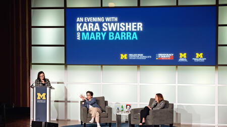 A photo of Kara Swisher and Mary Barra sitting on the stage, with Andrea Forte standing behind a podium