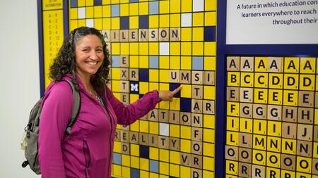 A photo of advisory board member Nikki Roda pointing to "UMSI" on a scrabble board display adhered to a wall