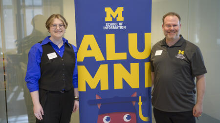 A student stands to the left of a "UMSI Alumni" banner, with their father standing to the right