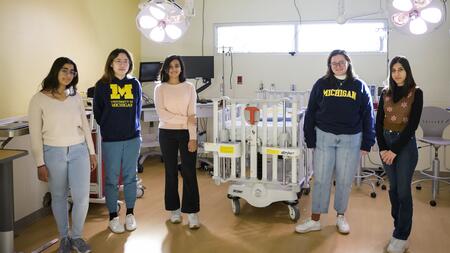 A photo of five students standing in a light-filled operating room. Two of them wear "University of Michigan" sweatshirts.
