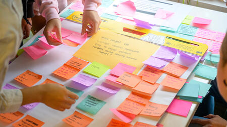 Bright multicolored post-its cover a white table, with a prompt visible in the middle: "How might we leverage common homeowner tools to engage upstream residents about reducing harmful runoff?"  