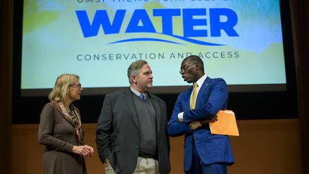 Elizabeth Yakel, Cliff Lampe and Lt. Gov. Garlin Gilchrist II stand on stage in front of a screen projecting "UMSI theme year 2023-24: Water conservation and access"