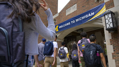 A photo of students wearing UMSI backpacks walking toward an arch with a University of Michigan banner clearly displayed at the entrance