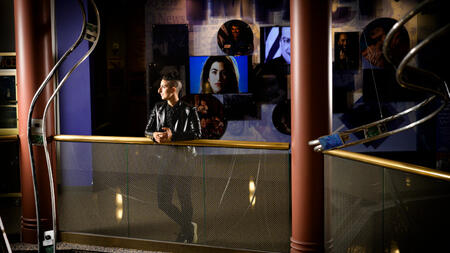 Tam Rayan wears a leather jacket and short hair and leans on a railing at the Arab American National Museum. 