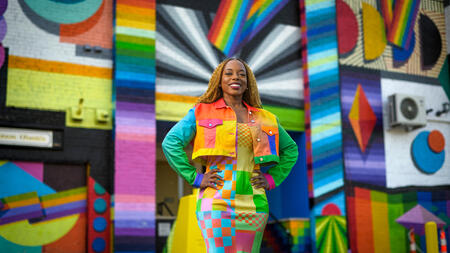 Megan Threats in a colorful jacket and a long skirt standing in front of a multi-colored wall. 