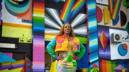Megan Threats in a colorful jacket and a long skirt standing in front of a multi-colored wall. 