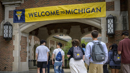 About 10 students walk into an outdoor brick passageway that is decorated with a maize and blue banner that reads "Welcome to Michigan." Two of the students wear UMSI-branded backpacks.