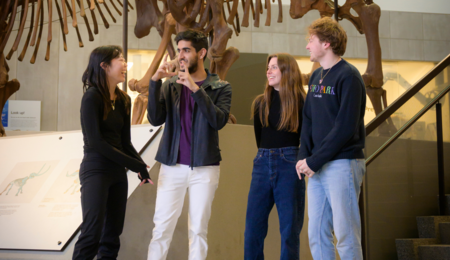 Four BSI students stand in a museum in front of a dinosaur skeleton, laughing and facing each other as one of them gestures to explain something