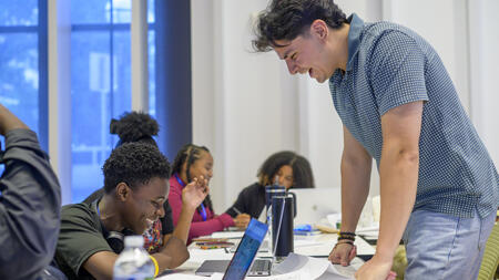 An education outreach assistant stands at a table facing a student who is smiling, holding a pen
