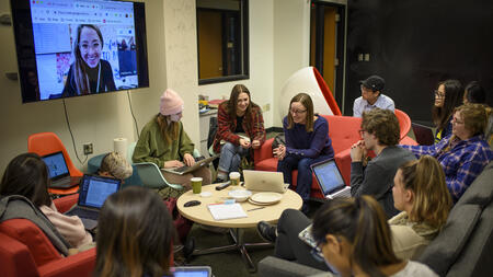 Gabriela Marcu sits in a circle with more than 10 students holding iPads. One student joins from a Zoom screen.