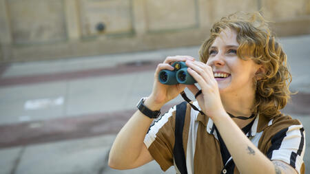 Meg Millure holds a pair of binoculars and smiles while glancing upward 