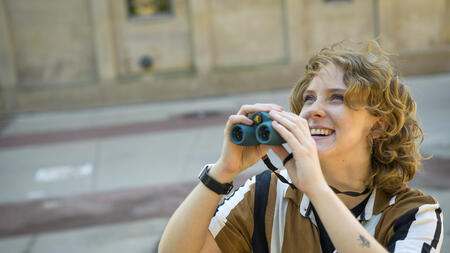 Meg Millure holds a pair of binoculars and smiles while glancing upward 