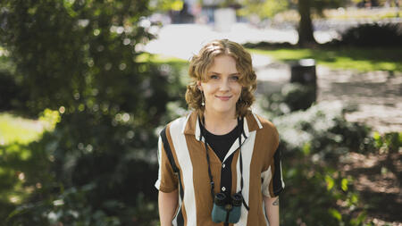 Meg Millure stands in a green area on campus with their hands in their pockets, wearing binoculars around their neck