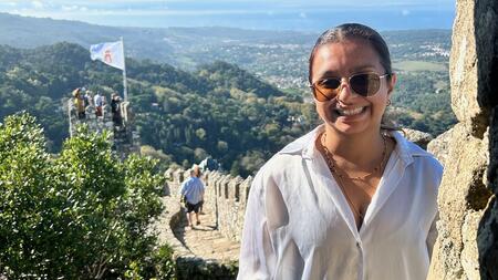 Jessica Thomas smiling standing on stone wall with turret and flag at its end. Background of rolling tree-covered mountains.