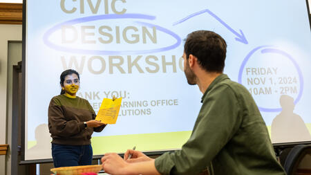 A student holds a piece of paper in front of a screen that reads Civic Design Workshop, while another student looks on