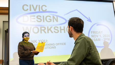 A student holds a piece of paper in front of a screen that reads Civic Design Workshop, while another student looks on