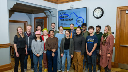 A group of sixteen participants and organizers from the civic workshop pose in front of a lobby monitor that says "Civic Design Workshop"