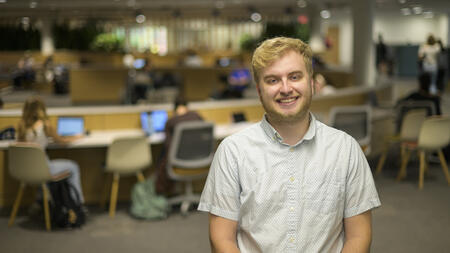 Austin Book poses in the library with out-of-focus students studying behind him