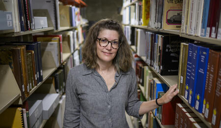 A librarian stands in the aisle between library shelves, with one arm resting on a book
