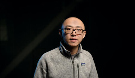 Jiayu Zhou wears a sweater and a black pair of glasses and looks at the camera.