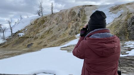 An individual in a red jacket, seen from behind, looks through binoculars at the snow-covered Mammoth Hot Springs