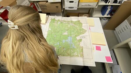 The back of a student's head as she studies a large map laid out on a table in front of her, with manuscript boxes visible on shelves in front of her