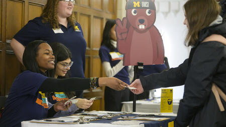 UMSI staff members sitting behind a table and handing stickers to students. Next to the table is a large cutout of a squirrel wearing a UMSI beanie.