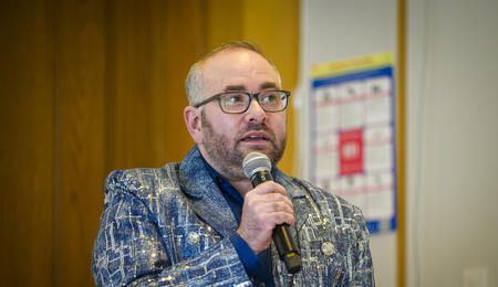 Oliver Haimson wearing a blue blazer and glasses and holding a microphone at his book launch. 