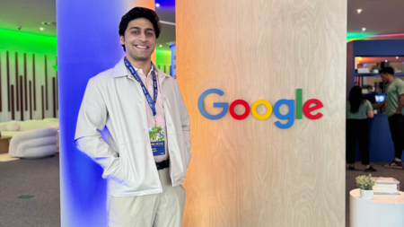 An individual smiles proudly wearing a lanyard and standing in front of a light wood wall adorned with the Google logo