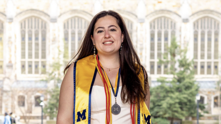A student wearing a maize stole, red cords, and a medal poses in front of an academic building with tall windows