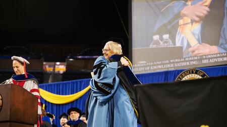 On stage in regalia, Elizabeth Yakel hugs a doctoral graduate. Someone stands at a podium to the left, and a screen and blue curtain are visible behind 