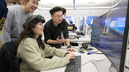 Students work together on code on a screen in a computer lab, one person is wearing a VR headset on their forehead.