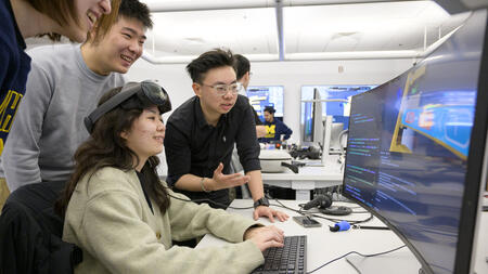 Students work together on code on a screen in a computer lab, one person is wearing a VR headset on their forehead.