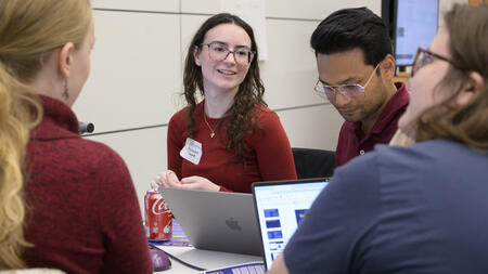 Four people sit at a table with laptops in front of them. One smiles while glancing into the group, as if in conversation