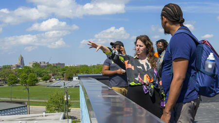 A person wearing a brightly patterned blouse stands on a rooftop with a few people gathered around, and gestures with one arm into the distance