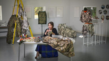 Sophia Brueckner poses with a small smile from a table inside a white-walled studio, sewing camo fabric, surrounded by camo fabric projects draped from a ladder and hanging racks