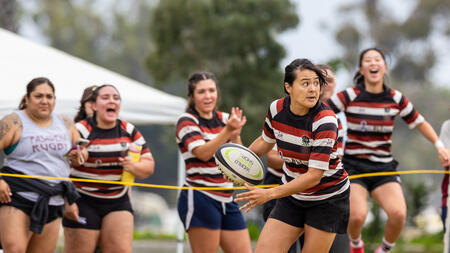 Action shot of Carolann Decasiano holding a rugby ball in the midst of a match, with teammates emoting on the sidelines behind her. They are dressed in matching striped uniforms.