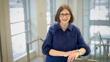 A portrait of Barb Smith in a dark blue blouse, leaning against a railing in the modern, sunlit Leinweber Building