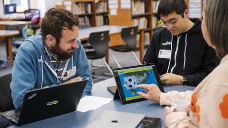 A person pressing a soccer icon overlaid on a world map on an iPad screen, as someone looks on and another person looks down taking notes