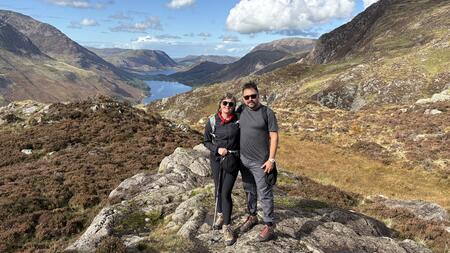 Katie McCurdy stands with another person in a rocky landscape with rolling hills, a body of water and a vast blue sky with billowing clouds stretching far behind them. They are both wearing sunglasses and hiking gear.