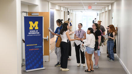 A group of students gather, facing each other and chatting, in a brightly lit hallway with a tall blue UMSI pull-up banner that says Information changes everything