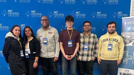 Simranjeet Singh poses with fellow hackathon participants in front of a backdrop with repeated MIT Hacking Medicine logos. Everyone wears nametag lanyards.