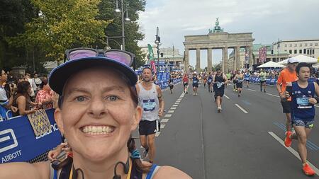 Lydia Schrandt smiling as she takes a selfie while running a race in a city amid other runners wearing race bibs. Onlookers cheer racers on from the sidelines.