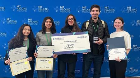 Gabriela Lagraba poses with fellow hackathon participants in front of a backdrop with repeated MIT Hacking Medicine logos. Lagraba and two others hold award certificates, and one person holds a giant check reading "1st Place Winner" made out to InStride for $2,000.