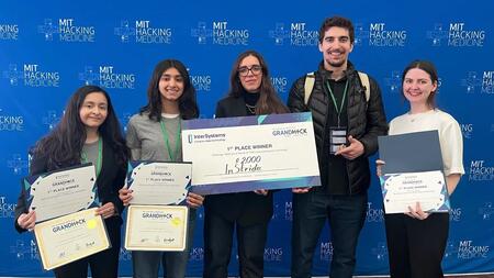Gabriela Lagraba poses with fellow hackathon participants in front of a backdrop with repeated MIT Hacking Medicine logos. Lagraba and two others hold award certificates, and one person holds a giant check reading "1st Place Winner" made out to InStride for $2,000.