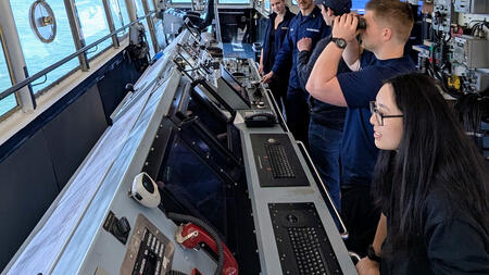 Students and U.S. Coast Guard personnel stand inside a vessel’s bridge, surrounded by navigation equipment and control panels, as one person looks through binoculars and others observe the water through large windows.