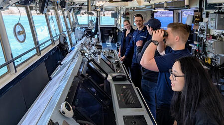 Students and U.S. Coast Guard personnel stand inside a vessel’s bridge, surrounded by navigation equipment and control panels, as one person looks through binoculars and others observe the water through large windows.