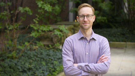 Card wearing glasses and a light purple button-down shirt standing outdoors with his arms crossed, looking at the camera.