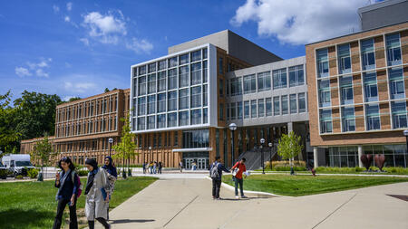 A far view of the Leinweber Building with scattered students walking the sidewalk in front of them, and two of them greeting each other by bumping fists